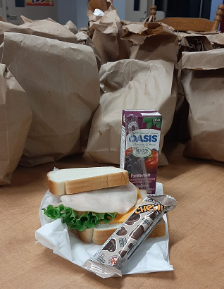 A sandwich, snack and drink, shown beside a stack of brown bagged lunches.
